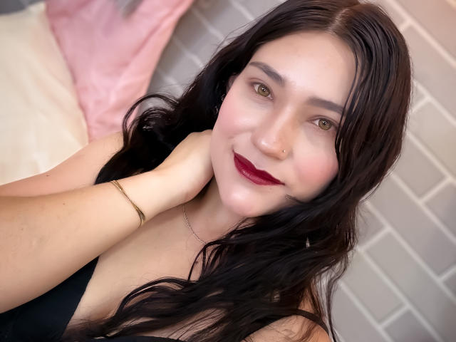 TamaraCians with long black hair, fair skin, green eyes, red lipstick, nose ring, black top, gold bracelet, smiling, hand on neck, soft light, brick wall background.