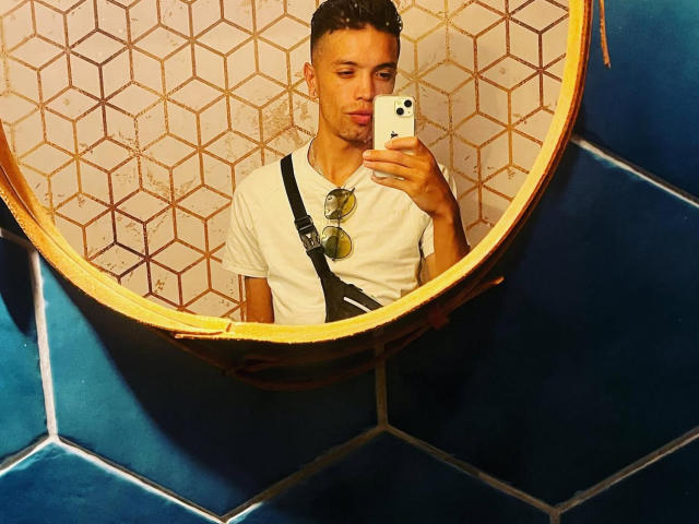 FinnDomHott, short curly brown hair, white t-shirt, black strap, sunglasses on chest, holding phone, mirror selfie, blue and hex-patterned wall, looking slightly pouty, handsome, casual.