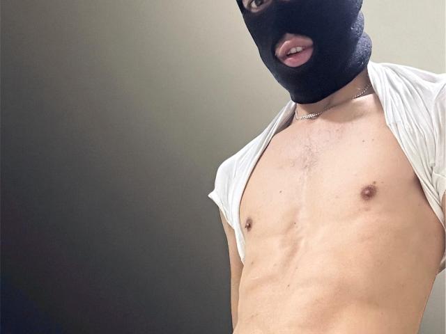 FinnDomHott with a black mask covering their head, tongue slightly out, white shirt pulled up, exposing their bare, slim, toned chest and flat, small ass.