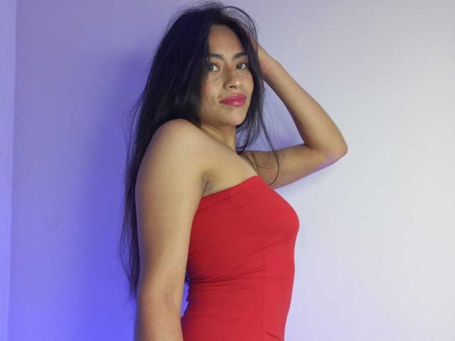 Emily Mitchell, avec ses longs cheveux noirs, sa robe rouge sans bretelles et son rouge à lèvres rose, se tient debout, un bras levé, montrant ses épaules nues et ses petites seins fermes..