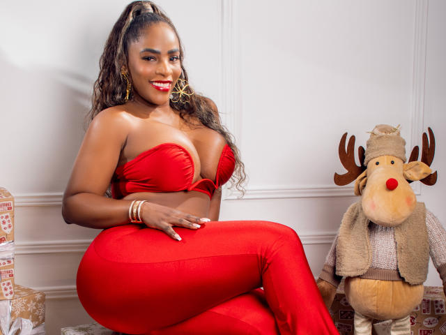 IsabellaKaned with large boobs in a red strapless top and red pants, sitting next to a reindeer stuffed toy. She's smiling, wearing gold earrings and a bracelet.