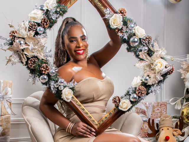 IsabellaKaned with dark skin, red lipstick, and long curly hair, wearing a beige dress, holding a Christmas wreath frame with pine, roses, and ornaments, smiling. Boobs, cute.