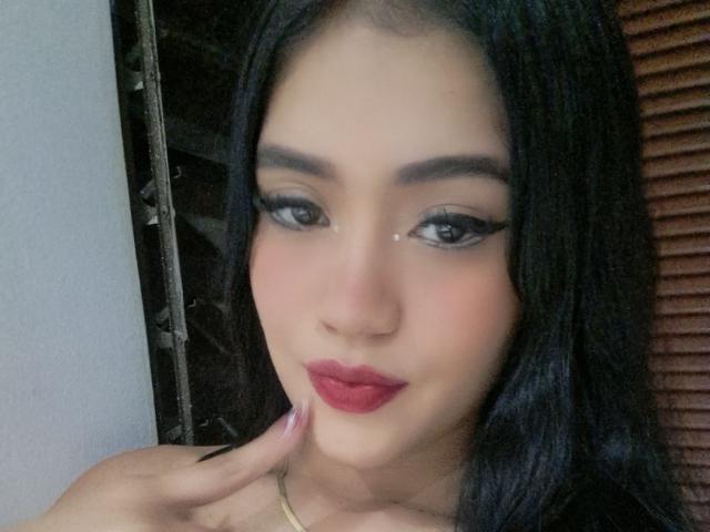 HottieSweettyHot with long black hair, brown eyes, and red lipstick, touching her chin. She has a tear under her right eye and is wearing a yellow necklace.
