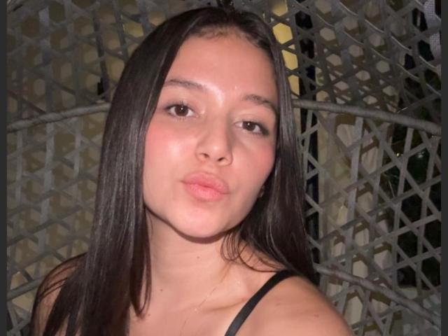 ZoeBarnett has long brown hair, a cute face, and is puckering her lips in a selfie. She's wearing a black spaghetti strap top, showing her small tits.
