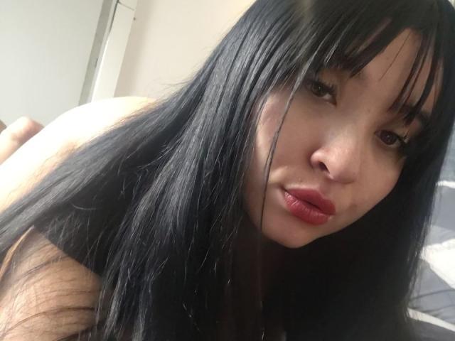LunaBellaHotty with long black hair, bangs, and red lipstick, looking at the camera with a slight pout, her bare shoulders and upper body visible.