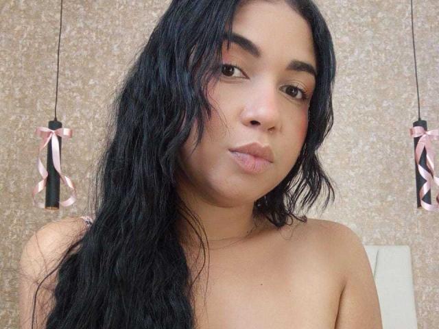 AliceSmity with long black wavy hair, brown eyes, and pinkish cheeks, pouting slightly, topless, showing her small, round tits against a textured beige wall.