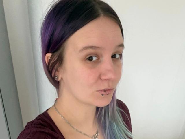 Sophiesoreal77-hot with purple and blue hair, nose piercing, lip piercing, and maroon shirt, wearing a chain necklace, has light skin and a neutral expression.