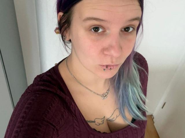 Sophiesoreal77-hot with purple and blue-dyed hair, nose piercing, lip ring, maroon shirt, necklace, and tattoos, looking up at the camera with a slight smile.