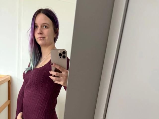 Sophiesoreal77-hot with purple and blue hair, wearing a maroon knit dress, taking a mirror selfie with an iPhone, showing her tits and a slight smile.