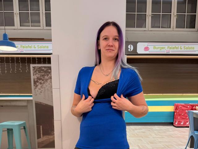 Sophiesoreal77-hot, with purple and black hair, wearing a blue dress, lifting it to reveal black lace-covered tits, standing in a white-walled, indoor space, smiling slightly.