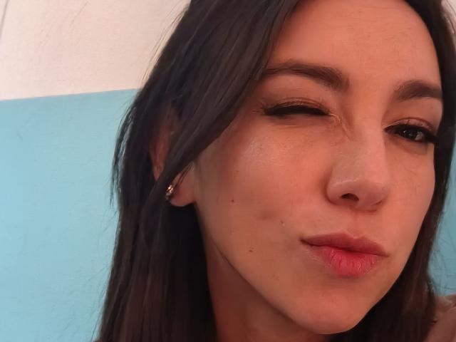 MiaReed with one eye closed, pouting lips, wearing small gold earrings, brown hair, light skin, slight freckles, and a subtle smile. Her nose is slightly upturned.