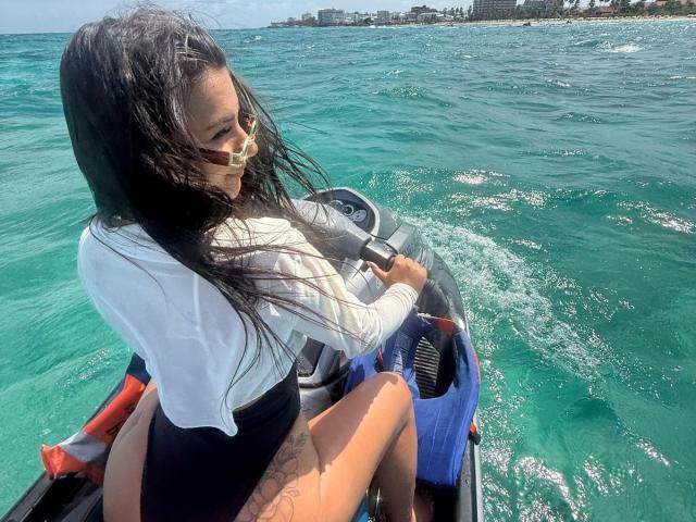 AlessiaFerry with wet black hair, white long-sleeve shirt, black shorts, riding a blue jet-ski, holding the handle, wearing yellow glasses, water splashing, sunny day.