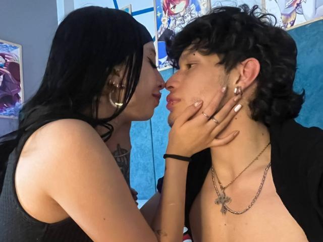 FuryDark with black hair and white nails kisses FuryDark's forehead, holding their chin. FuryDark has curly black hair, a chain necklace, and is shirtless. Boobs visible.