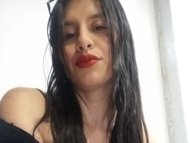 MadissonFontaine with wet black hair, red lips, and black strap dress, showing bare shoulders, looking down with a slight smile, no makeup on nose, white wall in background.