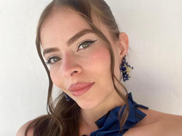 AnnaFer has light skin, green eyes with black eyeliner, brown hair in a ponytail, wearing blue ruffled top, and purple flower earrings. Her lips are pink and slightly glossy.