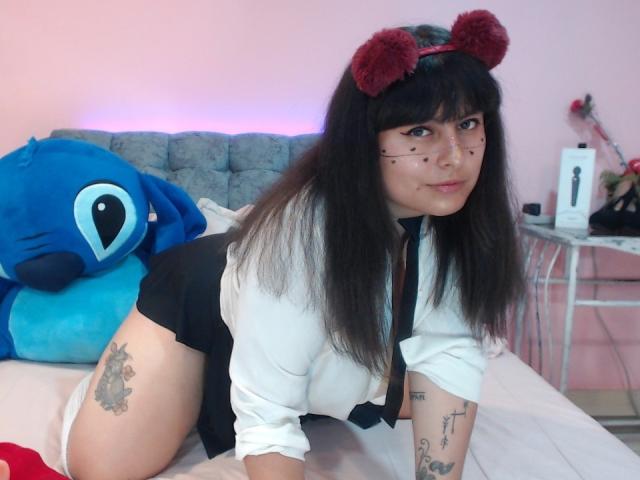SektethLilith on all fours, wearing a white shirt, black tie, and red pom-pom headband, with tattoos on her thigh and arm, and a blue plushie behind her.