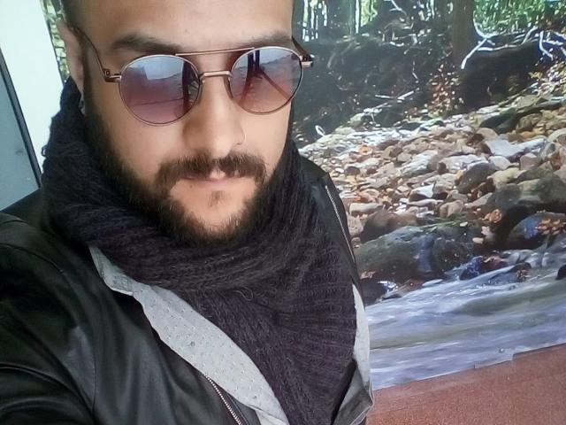 SektethLilith, com um bigode escuro, usando óculos de sol estilo aviador, um cachecol de malha preto e uma jaqueta de couro preta, tirando uma selfie. Um fundo com pedras visível..