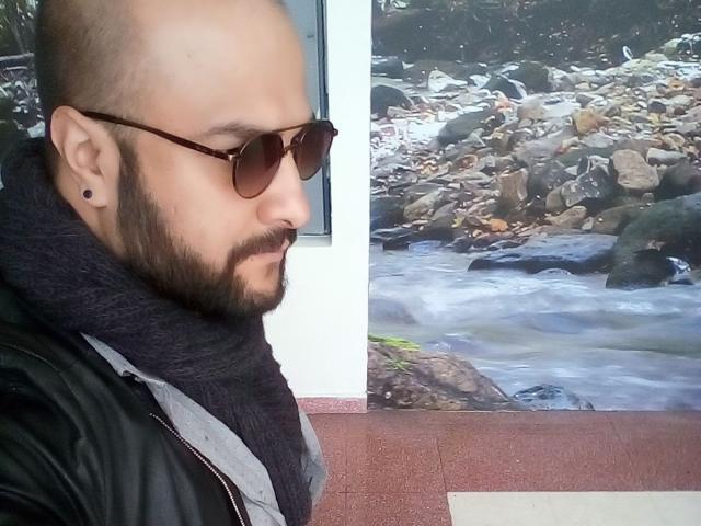 SektethLilith has a bald head, brown aviator sunglasses, dark beard, black scarf, and black jacket, standing by a rocky stream in a photo.