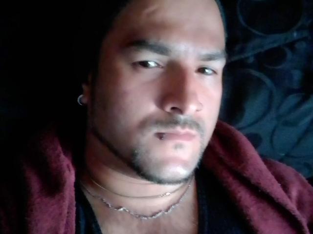 SektethLilith tem a cabeça raspada, um bigode escuro, barba e um piercing no nariz, vestindo uma toalha vermelha e uma camisa preta com uma corrente de prata..