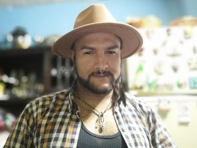 SektethLilith has a brown hat, plaid shirt, black tank top, and a necklace with a small pendant. He has a beard, nose ring, and a serious expression.