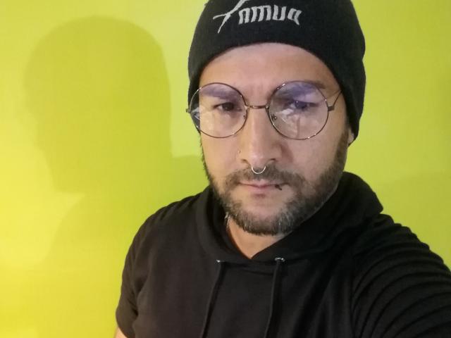 SektethLilith with glasses, black beanie, nose ring, and beard, wearing a black hoodie, has a serious face, and a yellow background. Slightly blurry selfie.