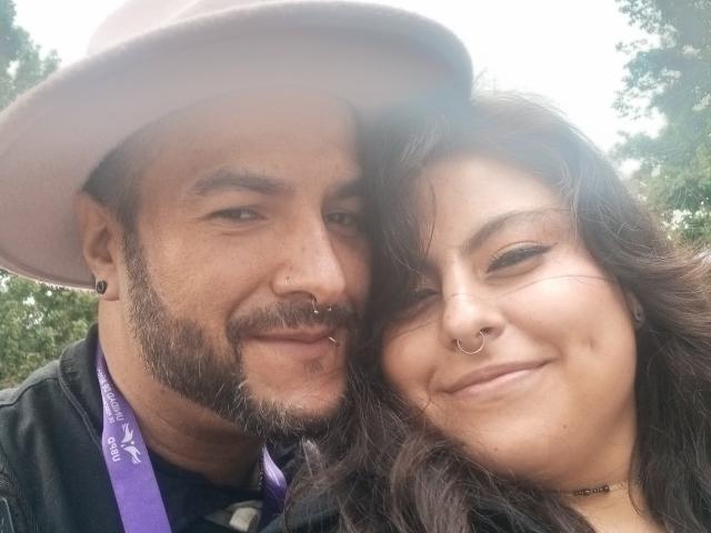 SektethLilith with a beard and purple hat, nose ring, and black jacket, next to SektethLilith with brown hair, nose ring, and choker, smiling. Both have visible tattoos.