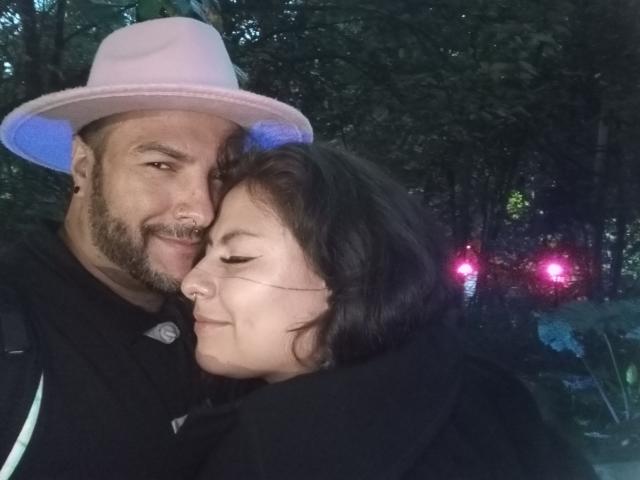 SektethLilith, a bearded dude with a pink hat, has his nose pierced and is hugging a brunette chick with closed eyes and a nose ring. Both are smiling.