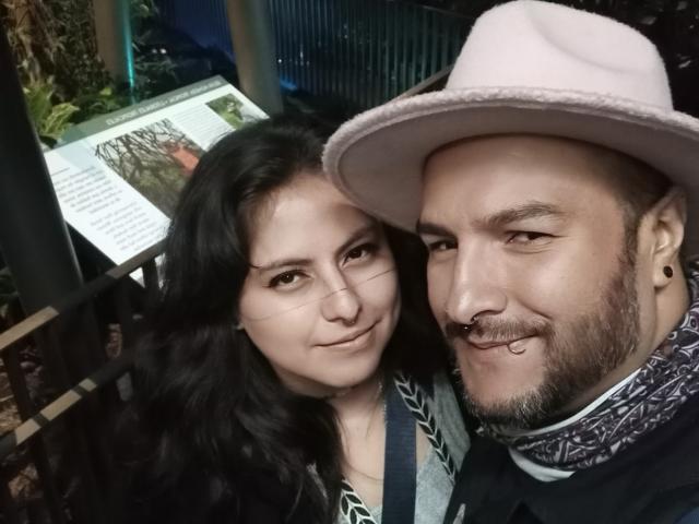 SektethLilith and a dude with a pink hat, nose ring, and beard, both smiling. She's got long black hair and he's got a patterned scarf. Slightly messy, cute.