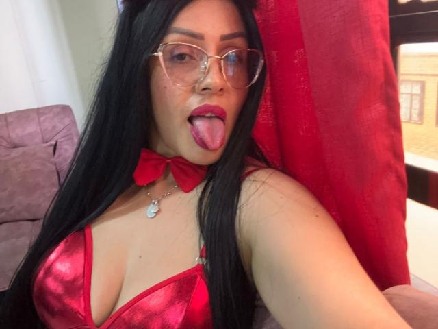 KinkyVane with long black hair, red glasses, and red bowtie, tongue out, wearing a shiny red bra, heart necklace, and red curtains, showing her boobs.