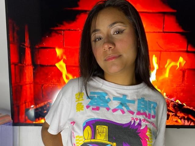 AnaRome with straight brown hair, light brown skin, wearing a white graphic tee, standing in front of a bright red fireplace, smiling slightly with a nose ring.