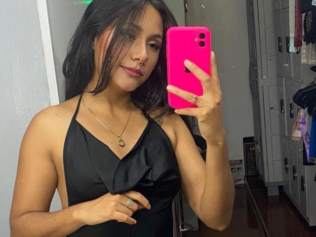 AnaRome with black halter dress, pink phone, nose ring, long dark hair, holding dress, gold necklace, ring, mirror selfie, showing tits. She's cute, confident, and sexy.