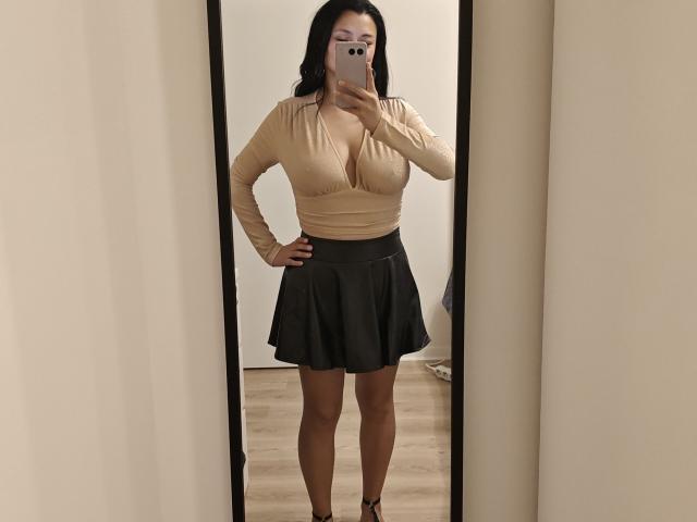 CharlotteGossart with big boobs in a beige top, black skirt, and black heels, taking a mirror selfie, holding a pink phone, standing on wooden floor.