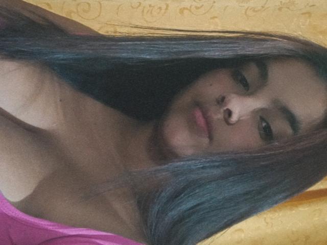 AmberMedellin, con cabello largo y oscuro, blusa rosa, pechos pequeños, una sonrisa encantadora, ojos marrones, piel de color marrón claro, mirando a la cámara, fondo de pared amarilla, iluminación suave, selfie casual..