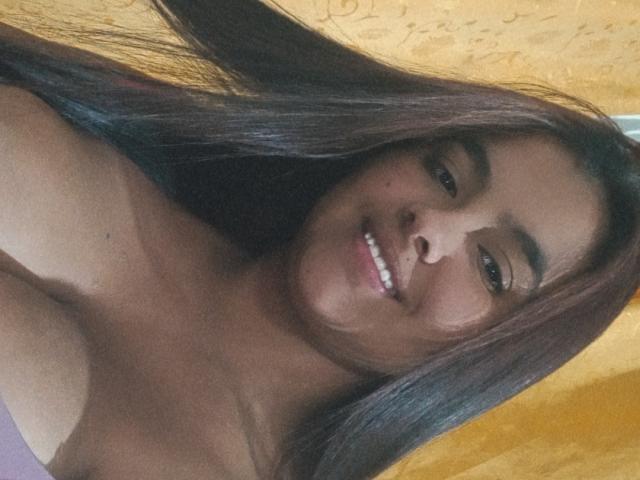 AmberMedellin mit langen, braunen Haaren, lächelnd, mit weißen Zähnen, und einem pinken Oberteil, das ihre nackten Schultern und den oberen Brustbereich zeigt. Ihre gebräunte Haut und ihr süßes Gesicht sind sichtbar..