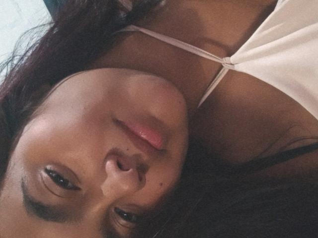 AmberMedellin's face, lying down, with dark brown hair, pink lips, and a white bra, showing her small tits and dark skin. Her eyes are looking at the camera.