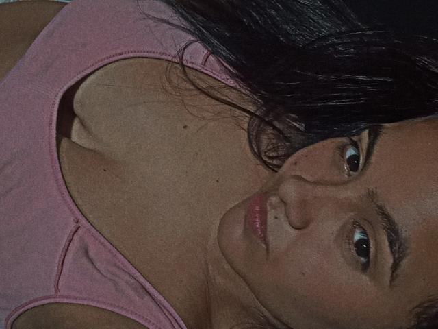Het gezicht van AmberMedellin is in beeld, ze draagt een roze tanktop, haar donkere haar valt over haar schouder, ze kijkt naar de camera en toont haar blote borsten en de decolleté..