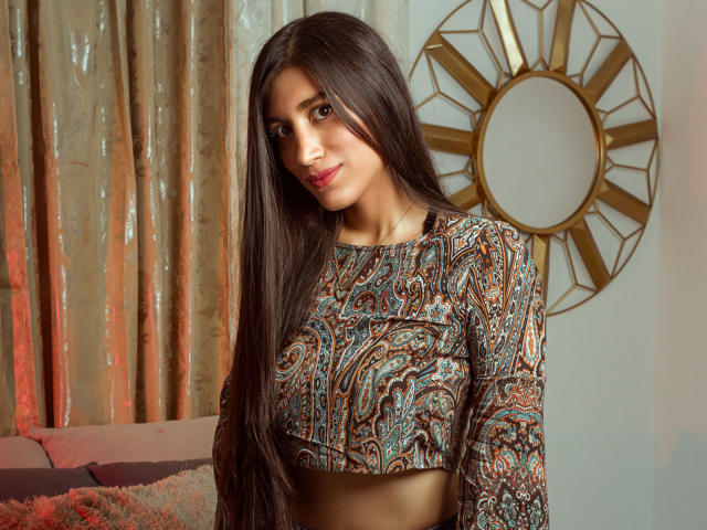 MaraClara with long dark brown hair, patterned crop top, showing her small tits, standing in a room with a gold clock and pinkish curtains. She has a cute smile.