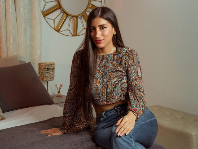 MaraClara with long brown hair, wearing a paisley crop top and blue jeans, sitting on a bed, her hands on the mattress, showing her ass in the photo.