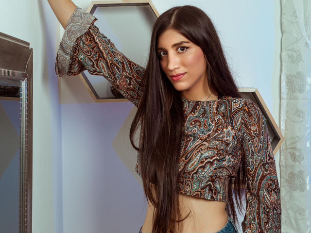 MaraClara with long brown hair, wearing a paisley crop top, arm raised, showing her tits, smiling slightly, standing indoors, blue jeans, light skin, brown eyes, and pink lipstick.