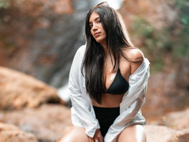 MaraClara with long black hair, wearing a black bikini and an open white shirt, sitting on rocks, showing her tits and ass, looking sexy with a slight smile.