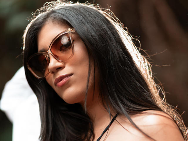 MaraClara with long black hair, brown oversized sunglasses, and a subtle smile, sunlight highlighting her tan skin and bare shoulders, looking cool and relaxed.