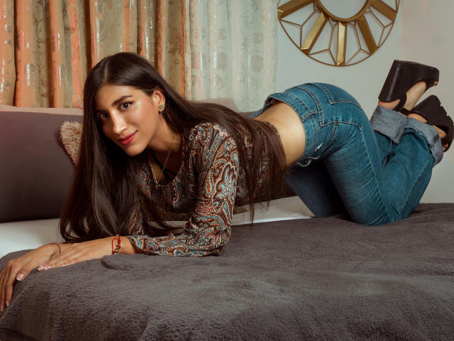 MaraClara with long brown hair, wearing a paisley shirt and blue jeans, on all fours on a gray bed, showing her ass, in black heels, smiling.