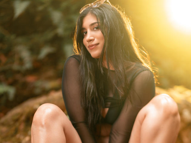 MaraClara with long black hair, wearing a sheer black top, sitting with knees up, sunlit, smiling, eyewear on head, warm light, soft focus, attractive, cute, sexy.