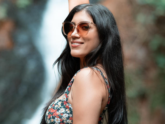 MaraClara with long black hair, wearing floral dress, brown aviator sunglasses, and smiling, has her arm raised, showing her cute nose and full lips, looking sexy.