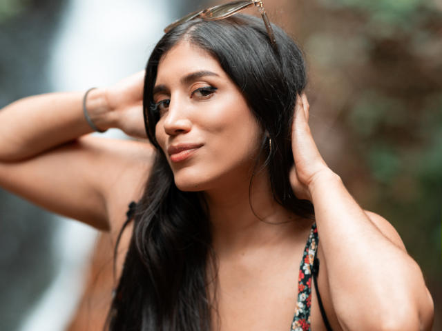 MaraClara with long black hair, tan skin, and subtle makeup, wearing a floral top, hands in her hair, sunglasses on her head, smiling softly, brown eyes, cute and sexy.
