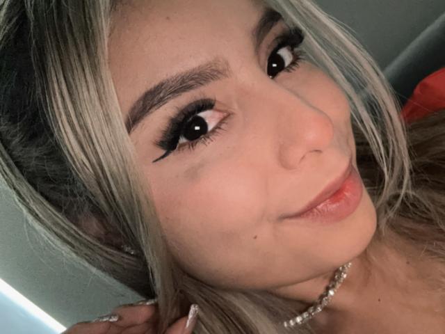 Amara Ricci mit blonden Haaren, schwarzem Eyeliner und rotem Lippenstift, lächelnd in die Kamera, trägt eine Perlenkette und sieht mit ihrer Hand auf dem Kopf niedlich aus..