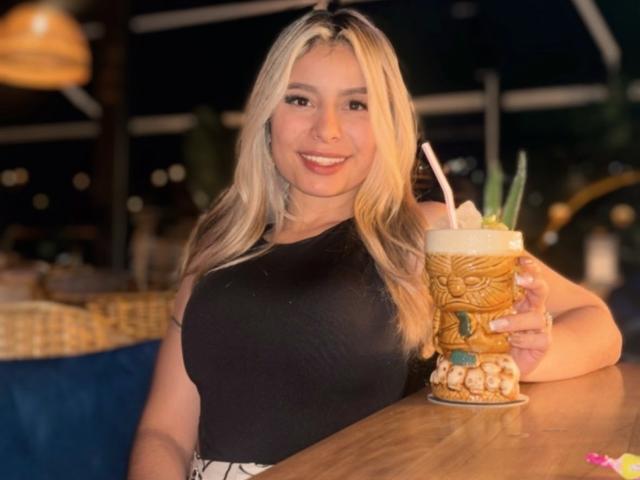 AmaraRicci with blonde hair, black sleeveless top, holding a tall, ornate drink with a straw, smiling, her big tits visible, in a dimly lit, cozy bar.