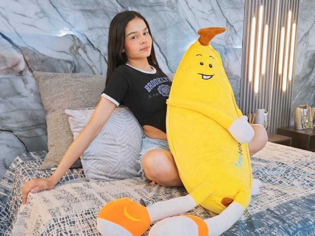 MiaGalvez sits on a bed with a yellow banana stuffed toy, wearing a black shirt and blue shorts, next to orange banana socks. She has long dark hair.