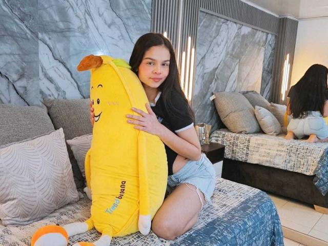 MiaGalvez with long dark hair, black shirt, and denim shorts, hugging a yellow Pikachu pillow on a bed, looking at the camera, in a modern, gray-themed room.