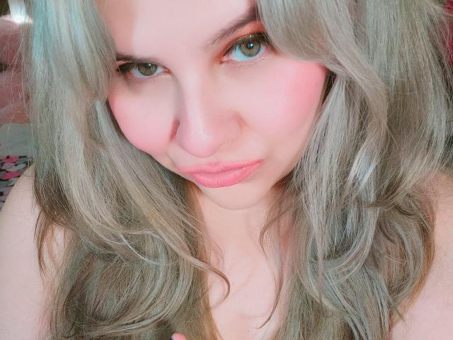 Akkanee has pale skin, pink blush, and pink lips, with long, wavy, silver-gray hair and green eyes, looking directly at the camera with a slight, cute smile.